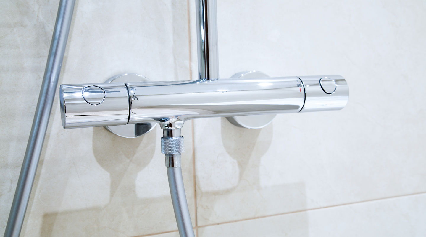 Gros plan sur colonne de douche thermostatique chromée avec deux molettes de réglage (température 38°C et débit), douchette à main et pommeau de tête, fixée sur carrelage blanc