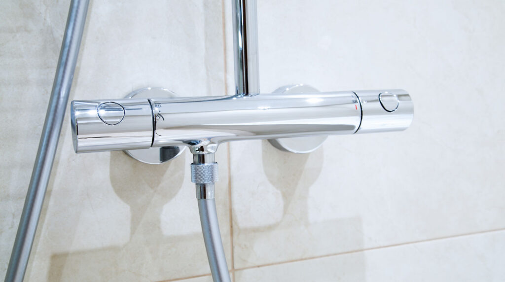 Gros plan sur colonne de douche thermostatique chromée avec deux molettes de réglage (température 38°C et débit), douchette à main et pommeau de tête, fixée sur carrelage blanc Gros plan sur colonne de douche thermostatique chromée avec deux molettes de réglage (température 38°C et débit), douchette à main et pommeau de tête, fixée sur carrelage blanc