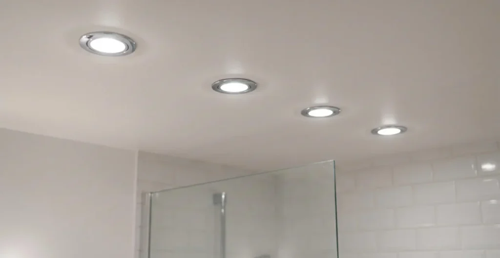 Vue en contre-plongée sur plafond blanc de salle de bain avec quatre spots LED encastrables ronds IP65 allumés, cerclage chromé, lumière blanche neutre, paroi de douche visible en arrière-plan Vue en contre-plongée sur plafond blanc de salle de bain avec quatre spots LED encastrables ronds IP65 allumés, cerclage chromé, lumière blanche neutre, paroi de douche visible en arrière-plan